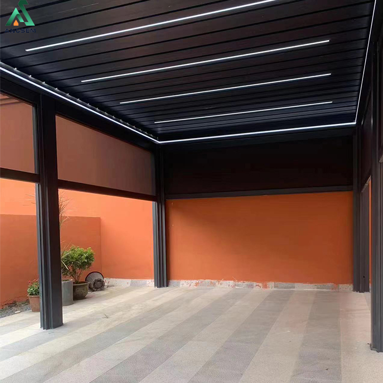 Interior of a black louvered pergola with windproof roller blinds partly lowered and integrated LED strip lighting in the louvered roof, set against an orange wall